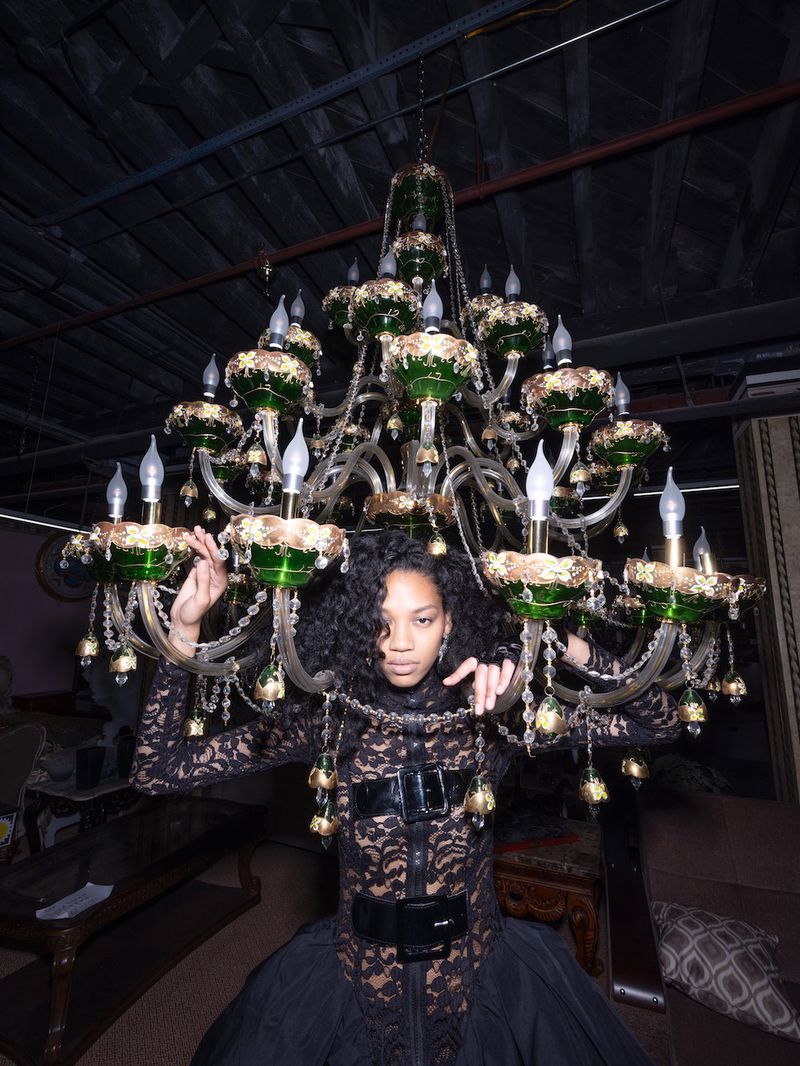 Woman in a black lace dress wearing a large decorative chandelier with lamps and green and gold accents in an indoor setting.
