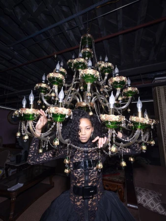 Woman in a black lace dress wearing a large decorative chandelier with lamps and green and gold accents in an indoor setting.