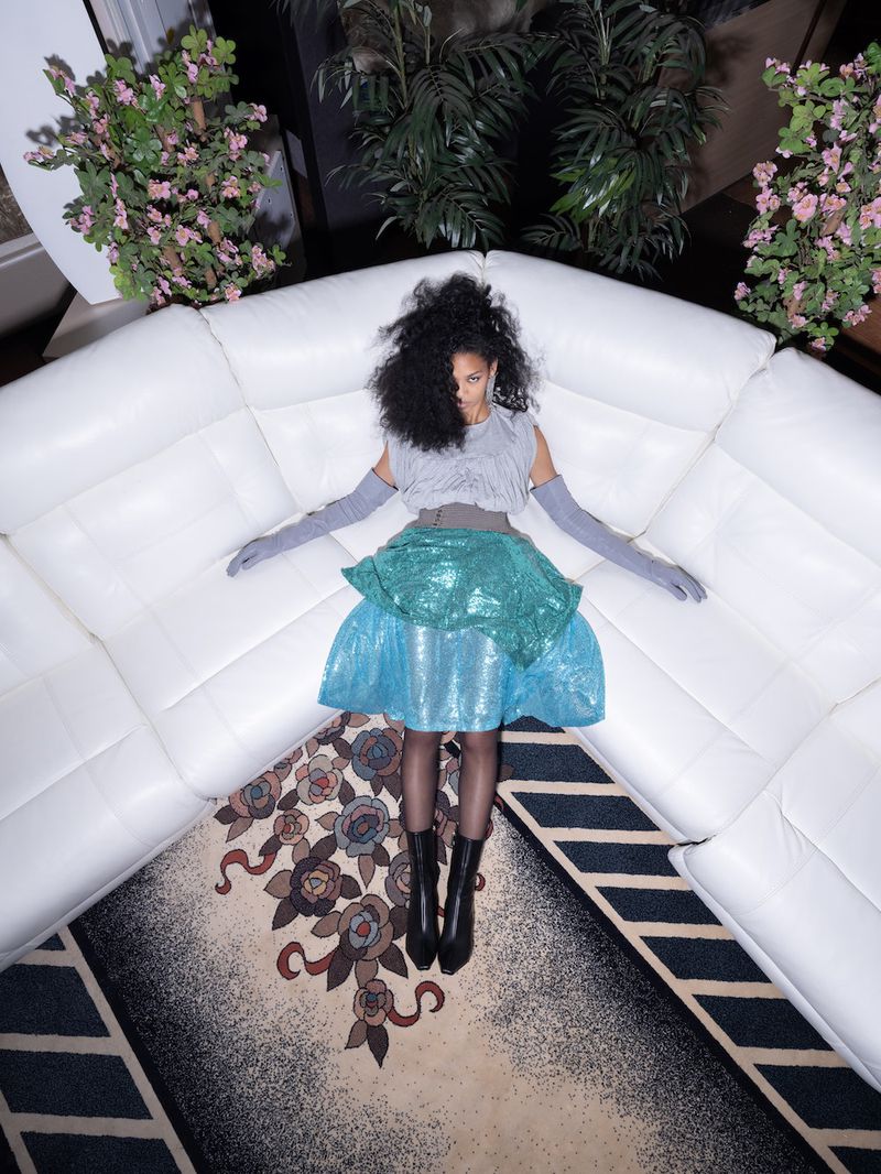 Overhead studio shot of a woman with dark curly hair reclining on a white corner sofa wearing a grey top and blue sequined skirt.