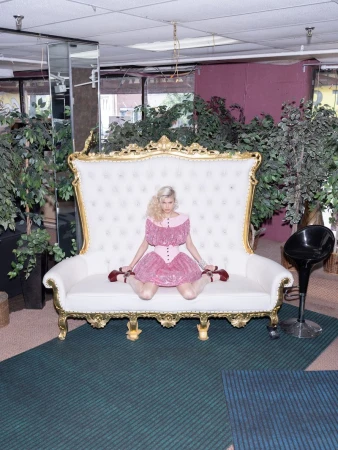 Blonde woman in a pink dress sits on an ornate white and gold sofa surrounded by potted plants, creating a vintage yet vibrant scene.