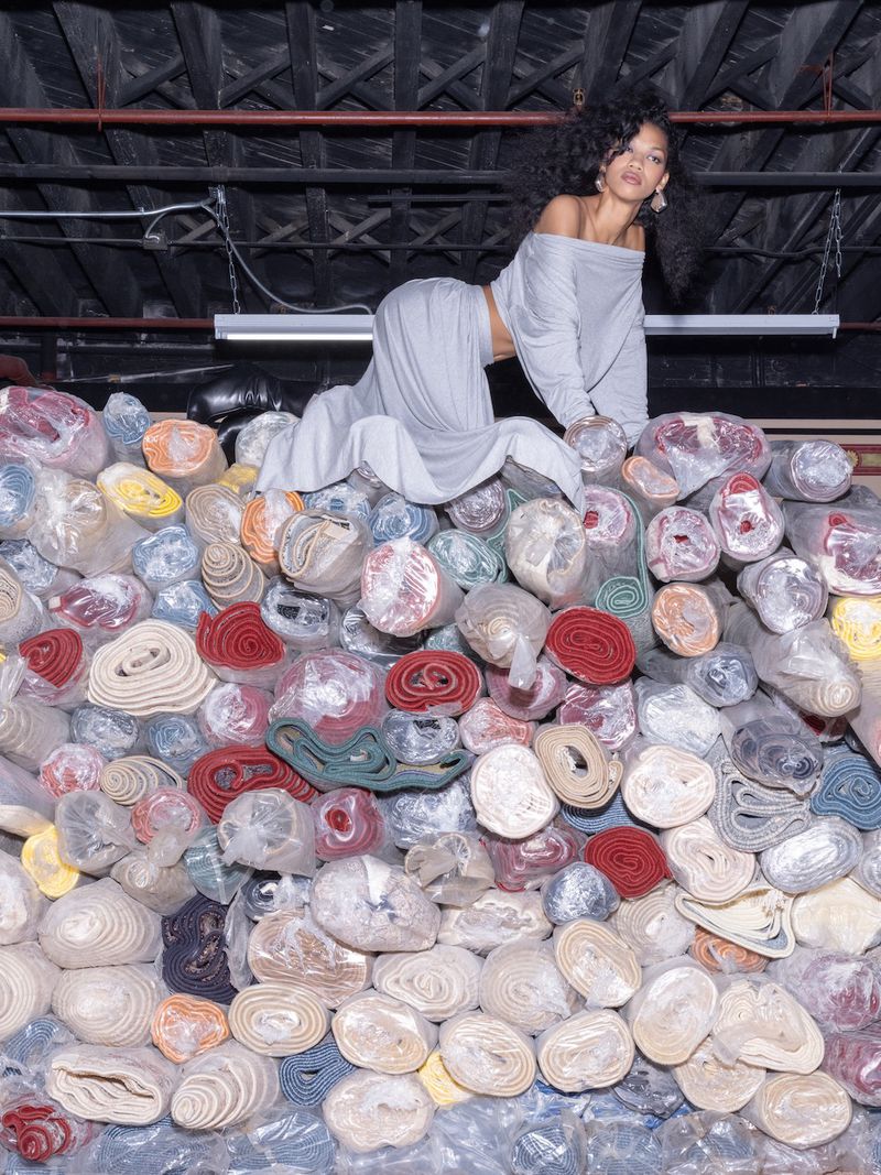 Woman in gray loungewear poses atop a colorful pile of carpet rolls in an indoor setting with dark wooden beams overhead.