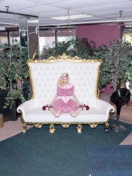 Model in a pink ruffled mini dress seated on an oversized ornate gold-framed white tufted throne sofa surrounded by potted plants.