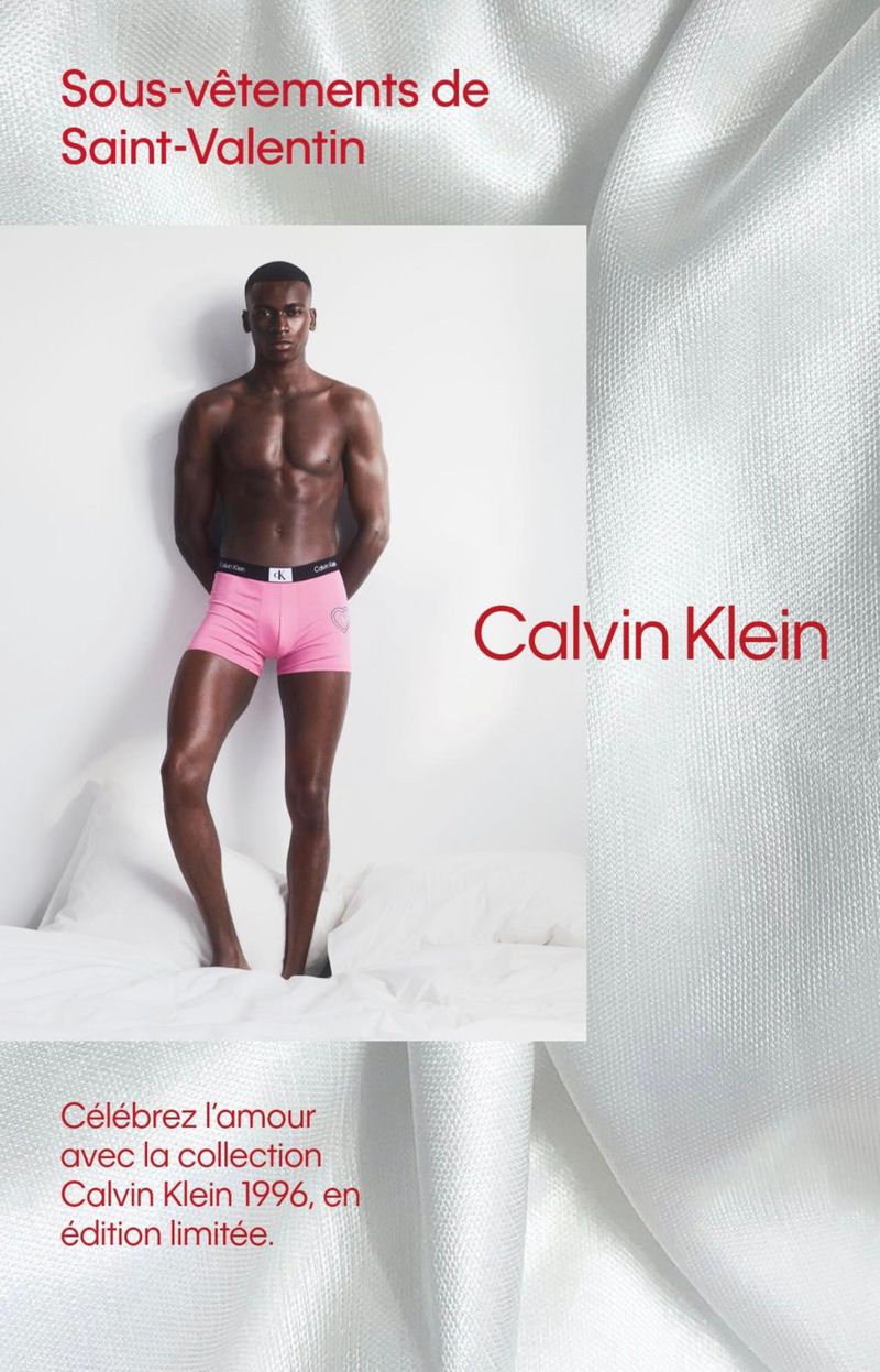 African American male poses in pink Calvin Klein underwear on white bed with white background for a Valentine's Day-themed advertisement.