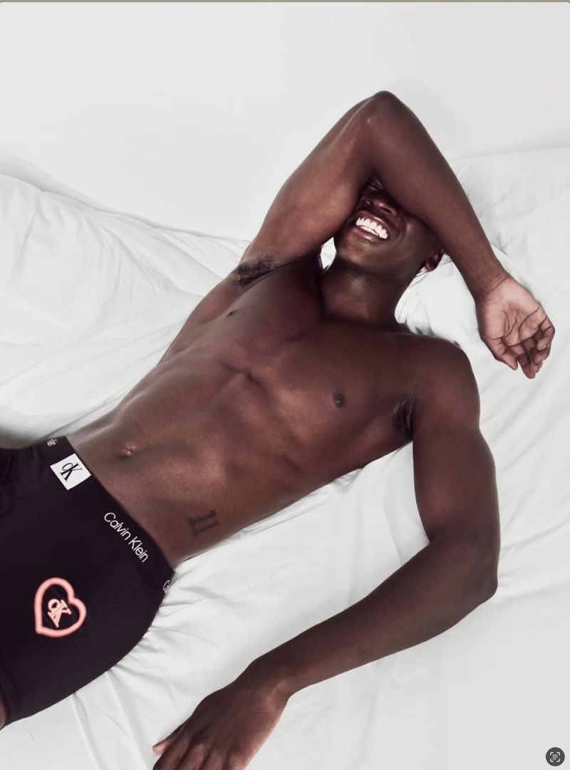 Dark-skinned adult man lying on a white sheet smiles with his arm over his face and wearing a Calvin Klein black boxer brief.