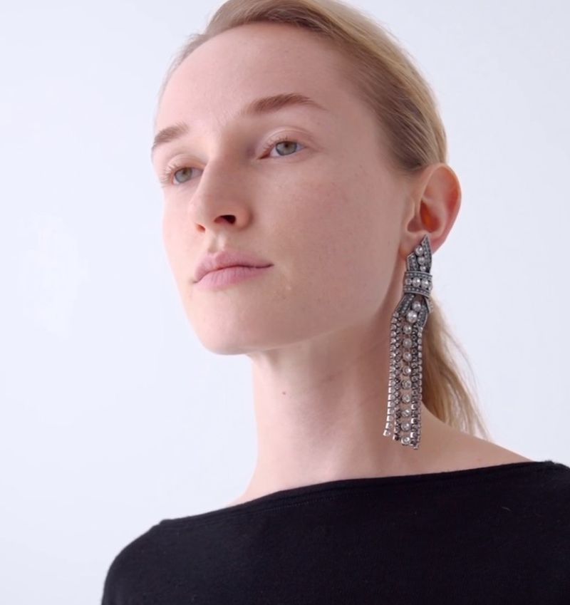 Woman with light skin and blond hair wearing large, jeweled dangling earrings against a plain white studio backdrop. Fashion Beauty shot.