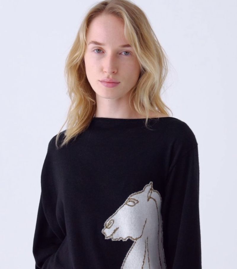 Blond woman in black long-sleeve shirt with white horse design against white background in medium studio shot, looking at camera.