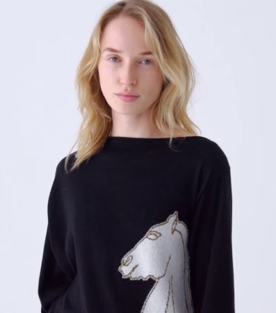 Blond woman in black long-sleeve shirt with white horse design against white background in medium studio shot, looking at camera.