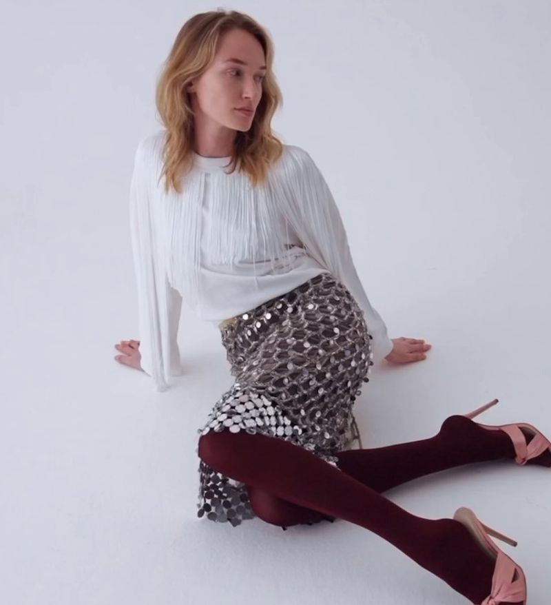 Fashion shot of a blonde woman sitting on the floor wearing a white fringe top and metallic skirt with maroon tights and high heels.