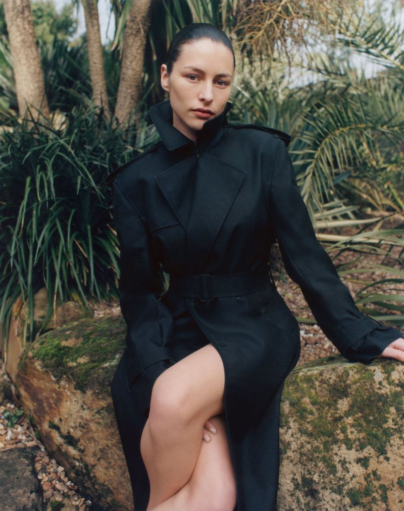 A woman poses outdoors on a mossy rock, wearing a long black belted trench coat, against a backdrop of green tropical foliage.