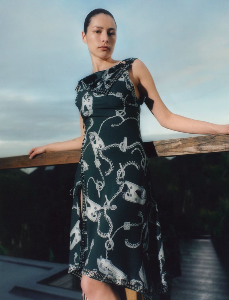 A woman in a patterned dress strikes a pose on a wooden boardwalk against a backdrop of soft blue sky and distant greenery.