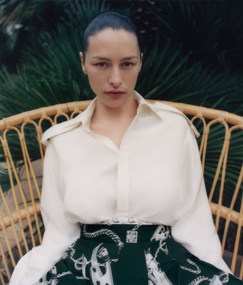 Elegant woman in a white blouse and printed skirt sits in a wicker chair against a leafy green background, portrait view.