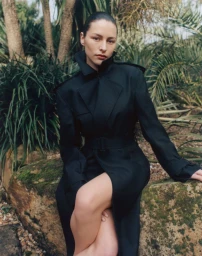 Model Phoebe Matthews seated on a rock wearing a black belted trench coat surrounded by garden foliage.