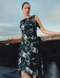 Model Phoebe Matthews in a printed sleeveless dark green dress leaning against a wooden balcony railing outdoors.