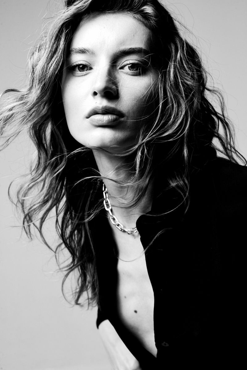 Monochrome portrait of a young woman with wavy hair wearing a dark, open collared shirt and silver chain necklace, neutral background.