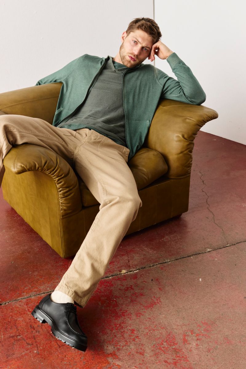 Man in green jacket and tan pants lounging in yellow armchair, black leather shoes, against red-and-white wall in studio.
