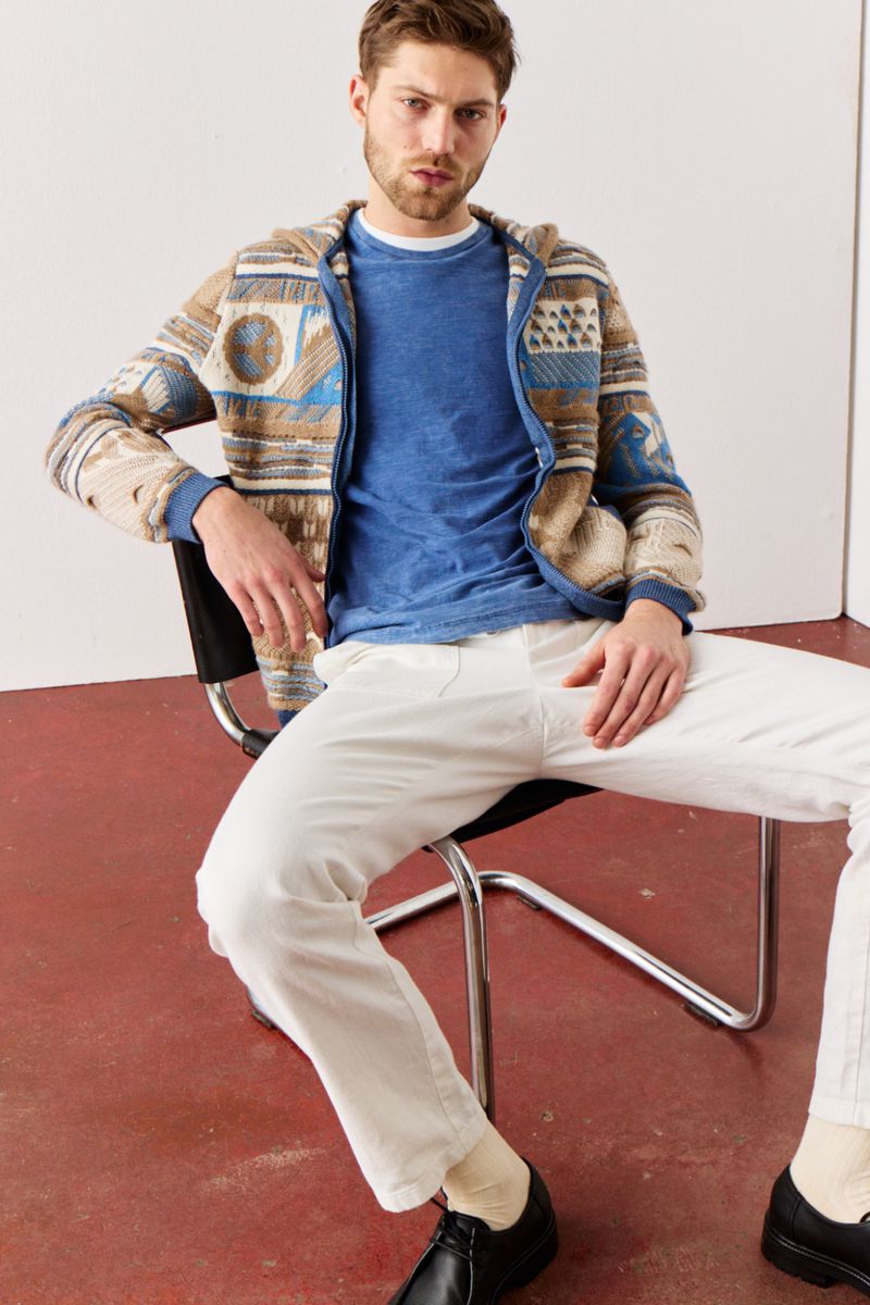 A man is sitting on a black chair. He is wearing a patterned hoodie, a blue t-shirt, and white jeans, posing in a brightly lit indoor space.