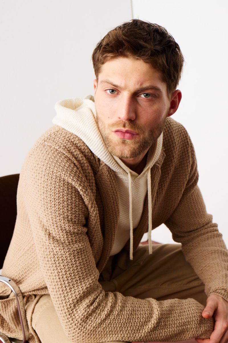 Portrait of an adult man wearing a hooded sweatshirt and a cardigan, sitting on a chair, against a neutral backdrop in a studio setting.