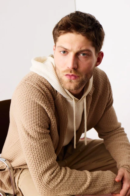 Portrait of an adult man wearing a hooded sweatshirt and a cardigan, sitting on a chair, against a neutral backdrop in a studio setting.