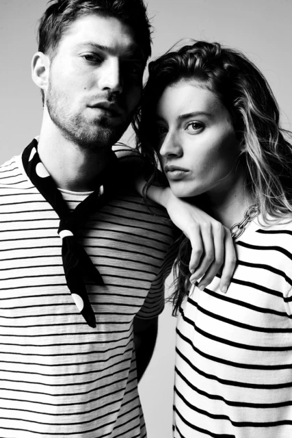 Black and white studio shot of a young adult couple in striped shirts. The man has a patterned scarf, and the woman wears a chain necklace.