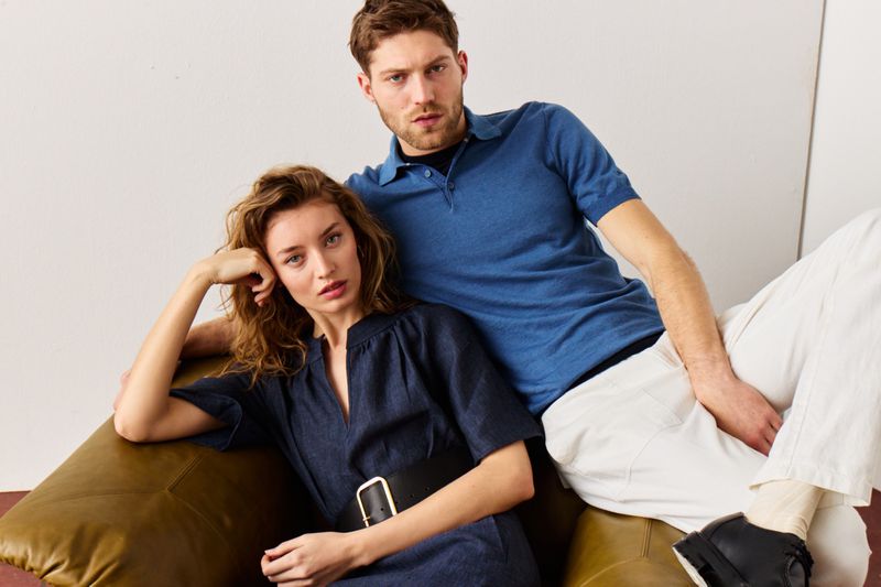 Attractive couple poses indoors wearing stylish matching blue and white clothing in a fashion editorial, seated on an olive-colored couch.