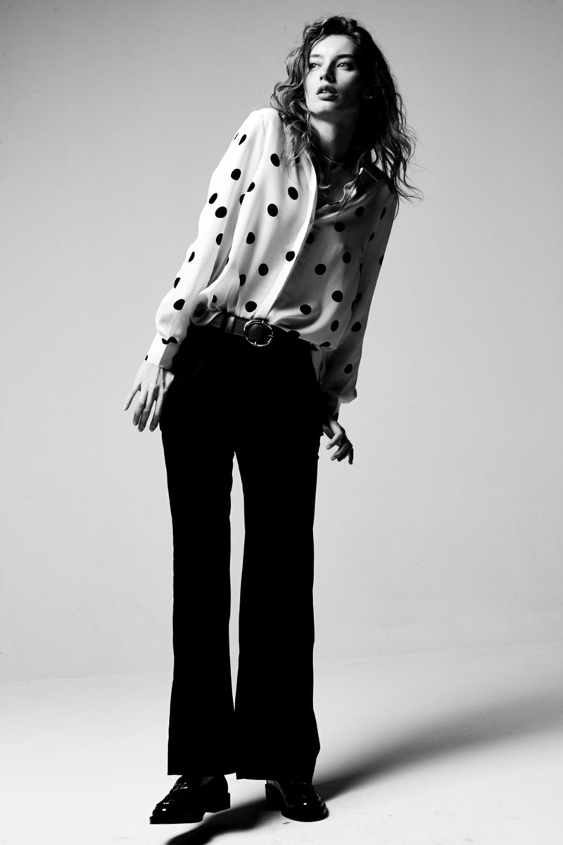 Monochrome full shot of a model with wavy hair wearing a polka dot blouse and black trousers against a neutral background in a studio.