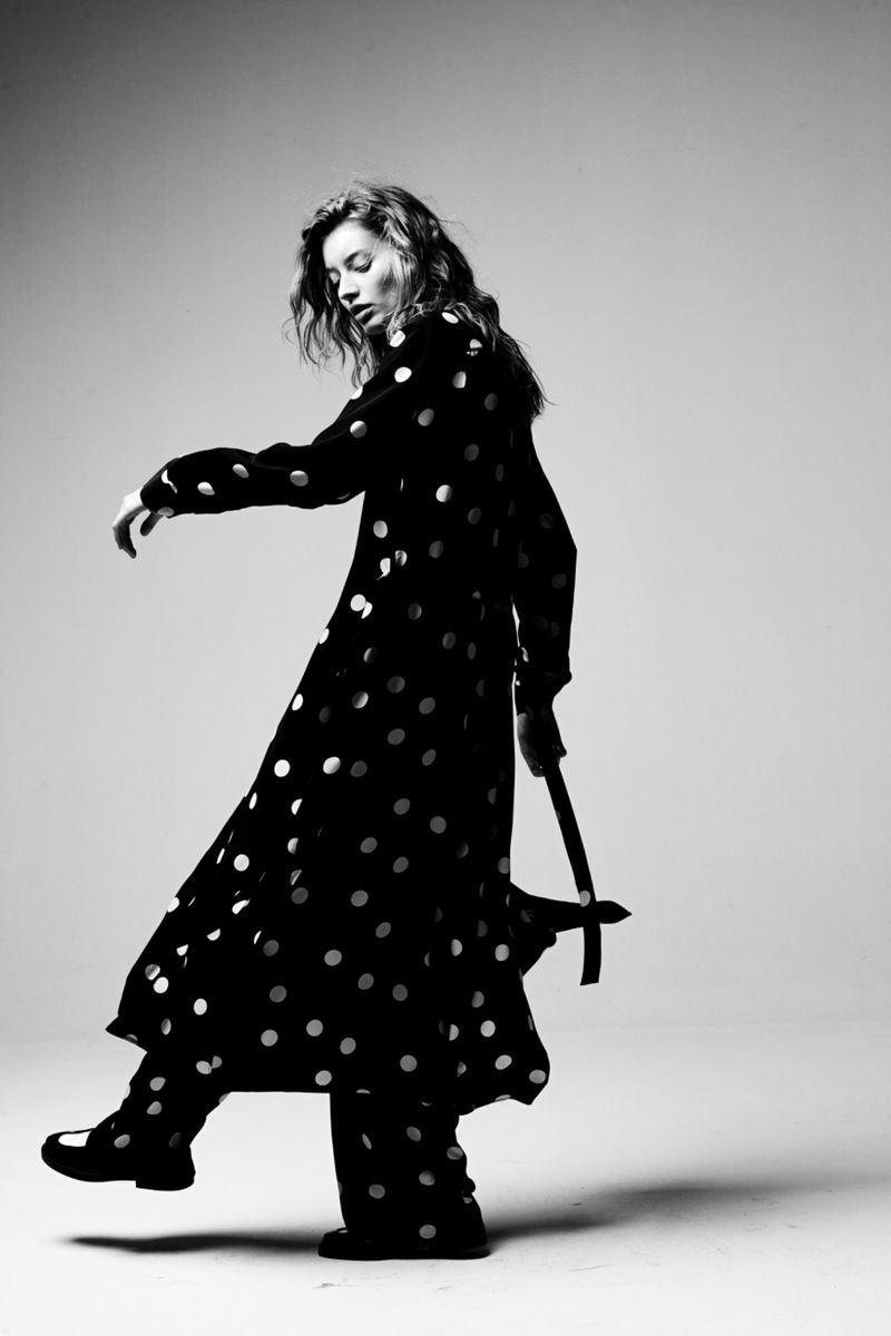 A woman with wavy hair poses in a black and white photo wearing a long-sleeved polka dot dress and dark shoes in a photo studio.