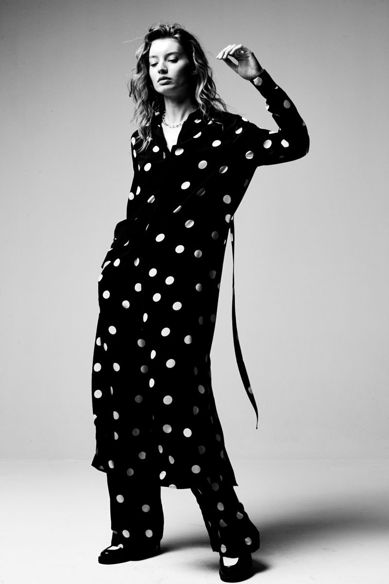 Monochrome fashion shot of an adult woman posing in a black and white polka dot shirt dress and pants in a photo studio setting.