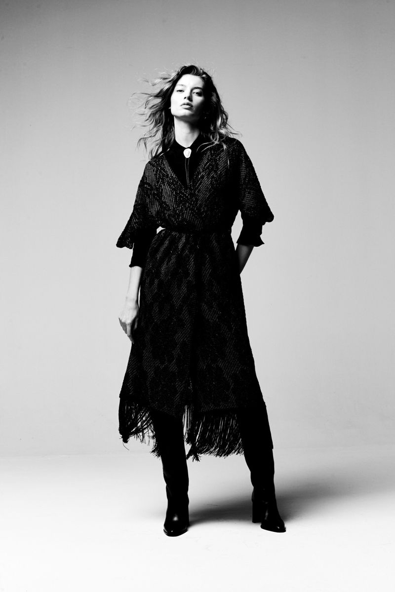 Fashionable woman in black dress and boots poses in studio, illuminated by bright lighting, creating an elegant black and white image.