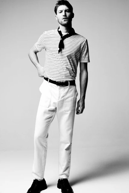 Black and white shot of a male model posing indoors in a striped t-shirt, white pants, black shoes, and a neck scarf against a white background.