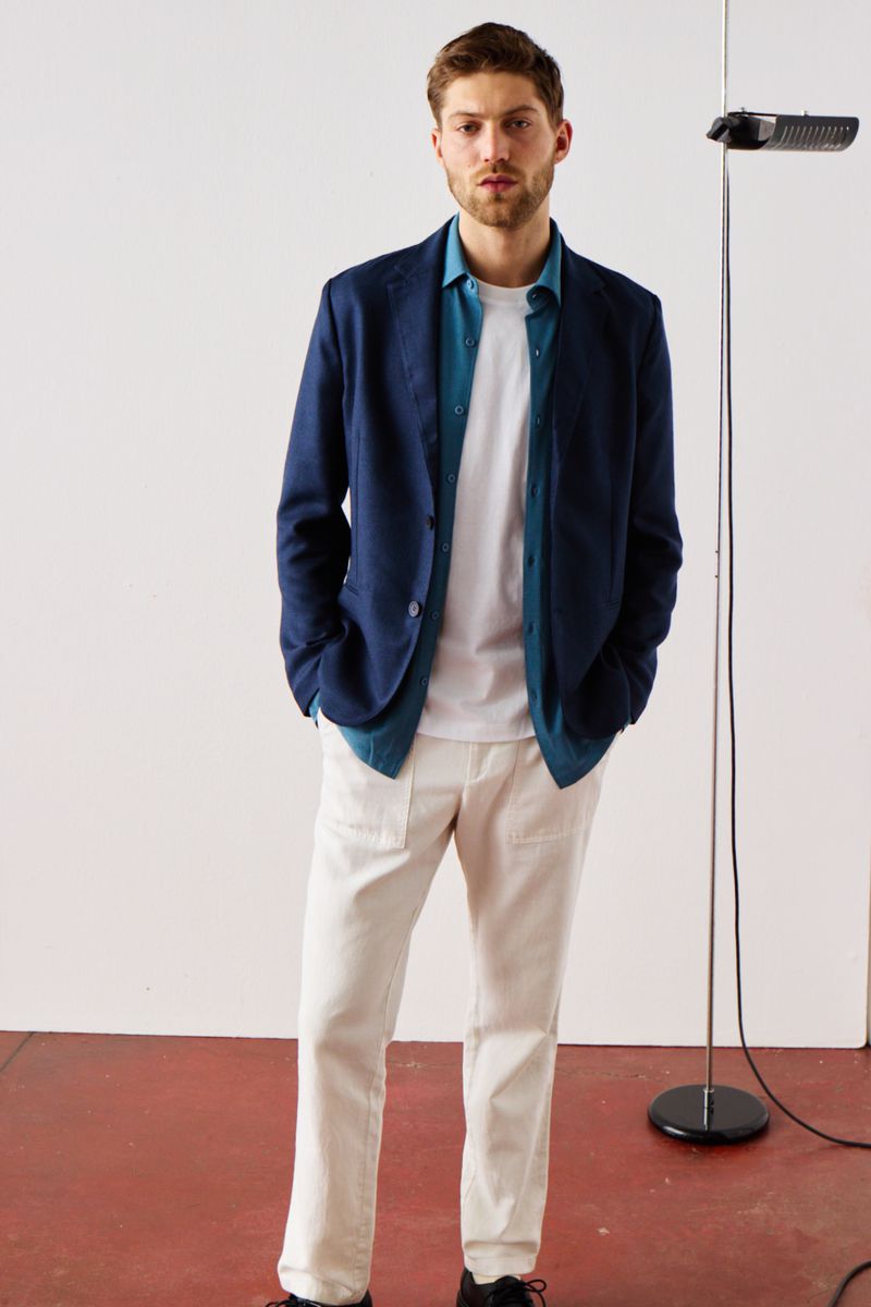 Full-length portrait of an adult man in a casual blazer, shirt, and trousers, standing in front of a plain white backdrop.