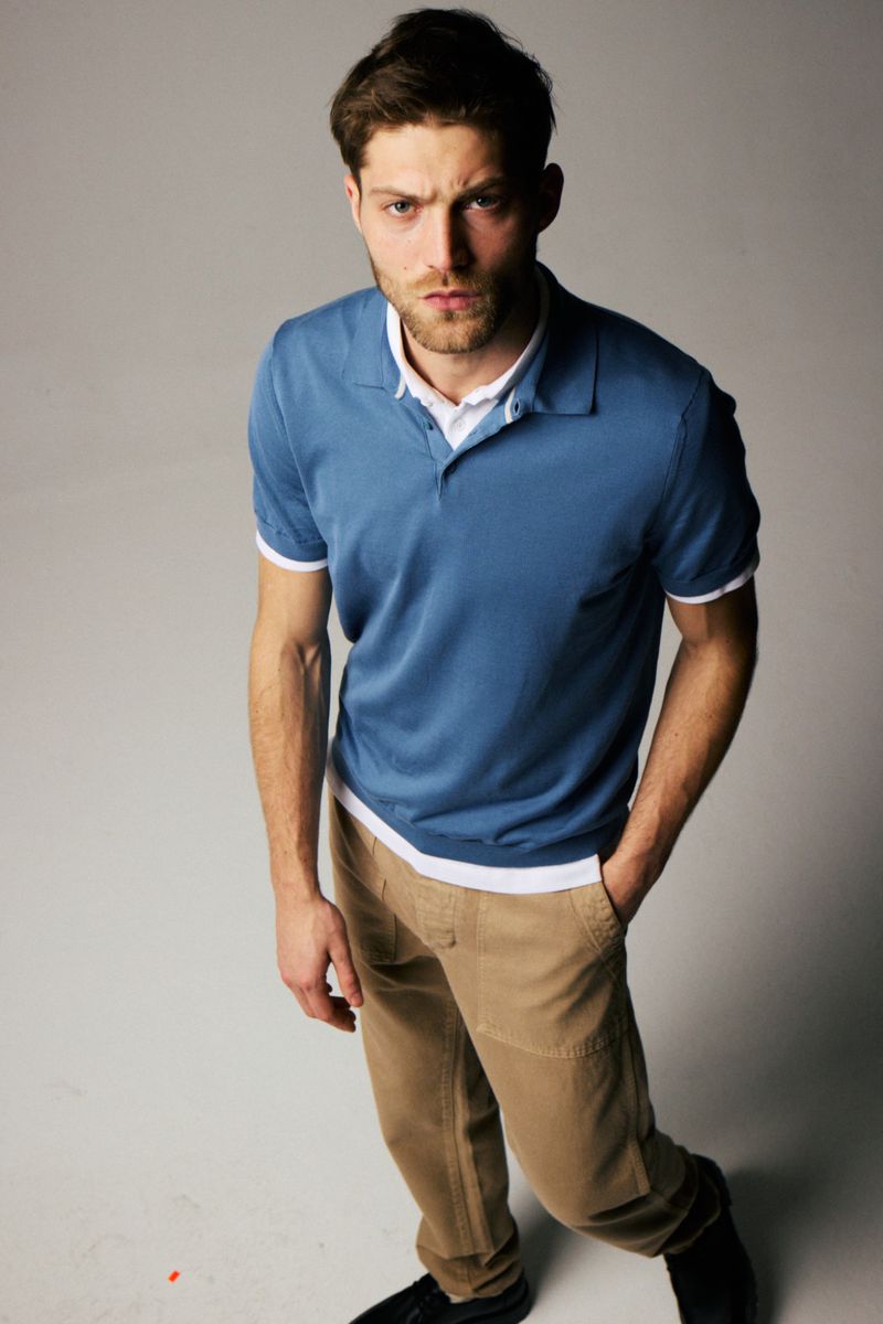 Studio portrait of a stylish adult male with a serious expression, wearing a blue polo shirt and tan pants against a plain background.
