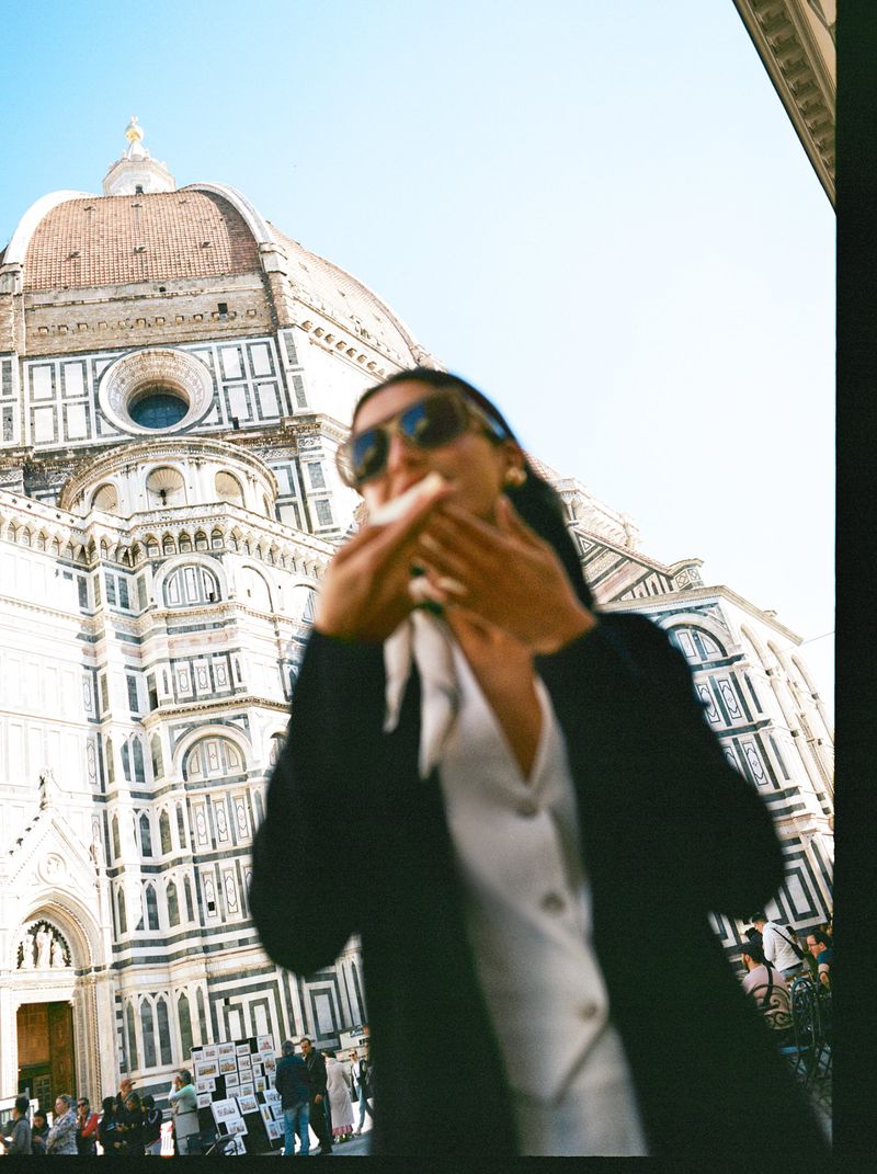 Woman in a blazer and sunglasses is eating in front of the Duomo in Florence on a sunny day with a clear blue sky.