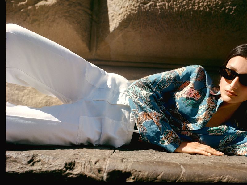 A young woman lies on stone steps, posing in white pants and a blue tiger-print shirt, wearing dark sunglasses in the sunlight.