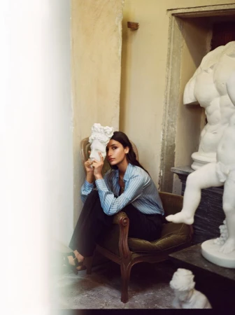 Indoor shot of a woman sitting on a chair, holding a white sculpture, with partial sculptures in the background in a neutral tone.