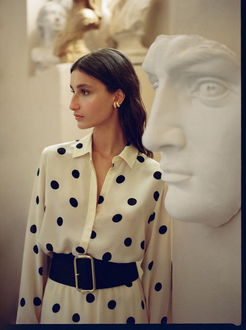 Young woman in a polka dot dress poses next to a classical bust in an art gallery, showcasing fashion and art in an indoor setting.