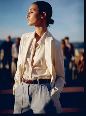 Model wearing an off-white blazer and silk button-down shirt outdoors in warm sunlight with eyes closed.