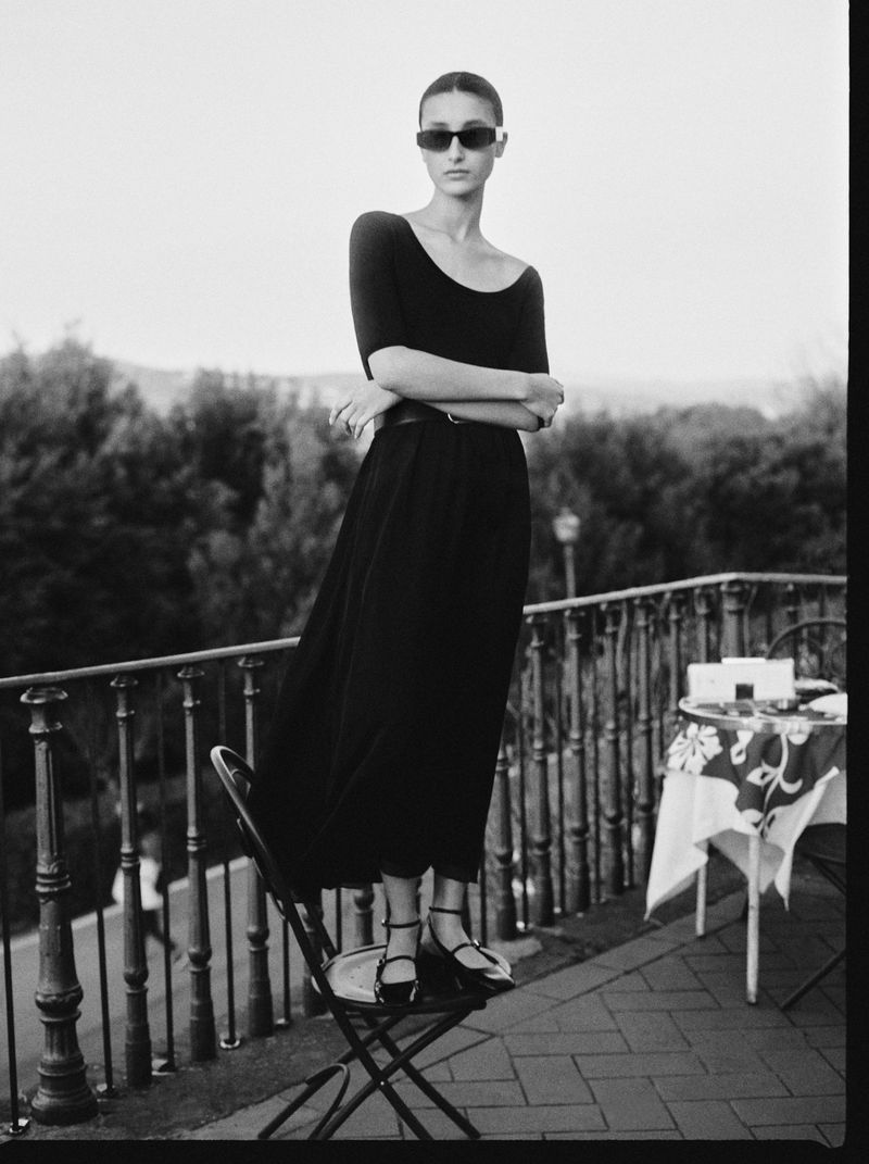 Elegant woman in sunglasses and a black dress stands on a chair on a balcony with a scenic view of trees in the background.