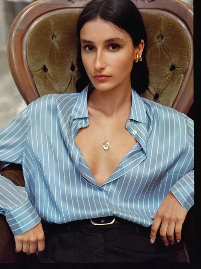 Model wearing a blue striped silk shirt and gold necklace seated in a tufted armchair for Falconeri.