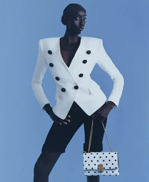 Fashion shot of a slender Black woman in a white jacket, black shorts, and a polka dot bag on a light blue backdrop.