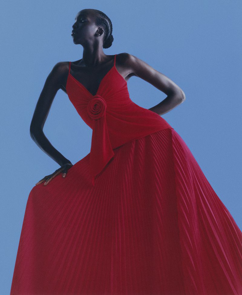 Woman in a red gown poses in front of a blue backdrop. The gown has thin straps and a rosette on the bodice, with a pleated skirt.