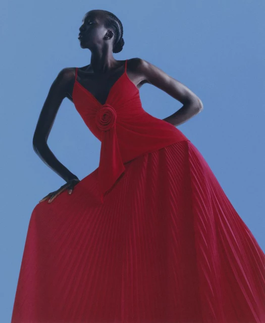 Woman in a red gown poses in front of a blue backdrop. The gown has thin straps and a rosette on the bodice, with a pleated skirt.