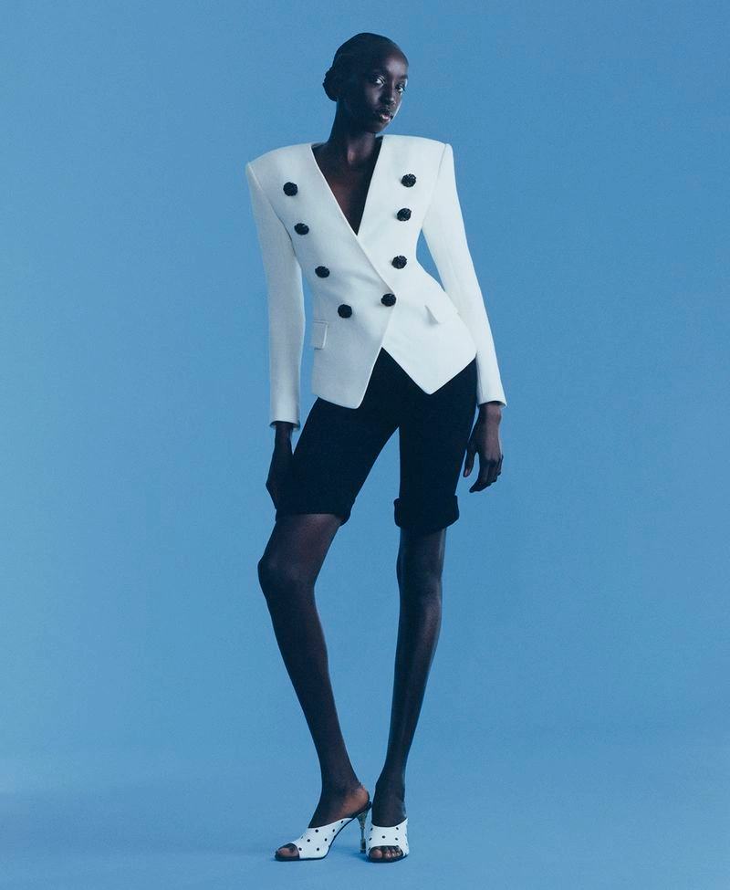 Model Nyaueth Riam wearing a structured white double-breasted blazer and black shorts against a blue seamless background
