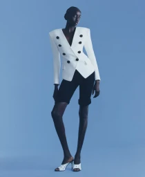 Model Nyaueth Riam wearing a structured white double-breasted blazer and black shorts against a blue seamless background
