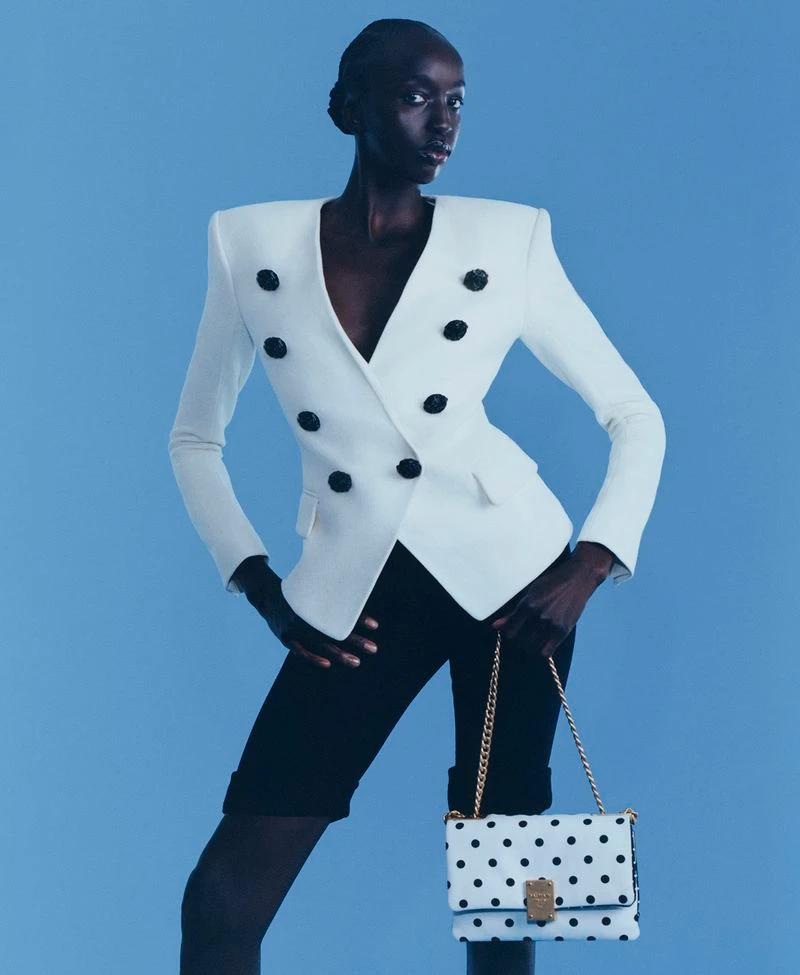 Model Nyaueth Riam poses in a white structured blazer and black shorts, holding a polka-dot handbag against a solid blue background.