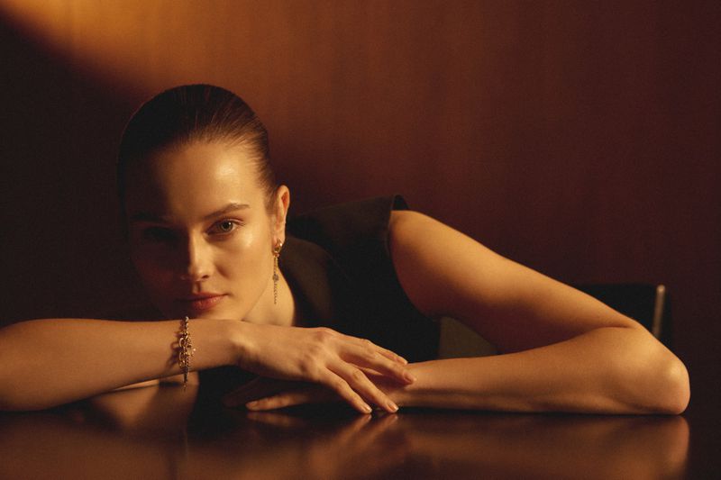 Woman in sleeveless top with bracelet and earrings rests her arms on a table in a warm, low-lit indoor setting.