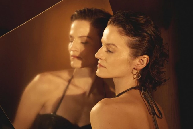 Model Jac Jagaciak in profile with gold hoop earrings and slicked back hair posing next to her mirror reflection.