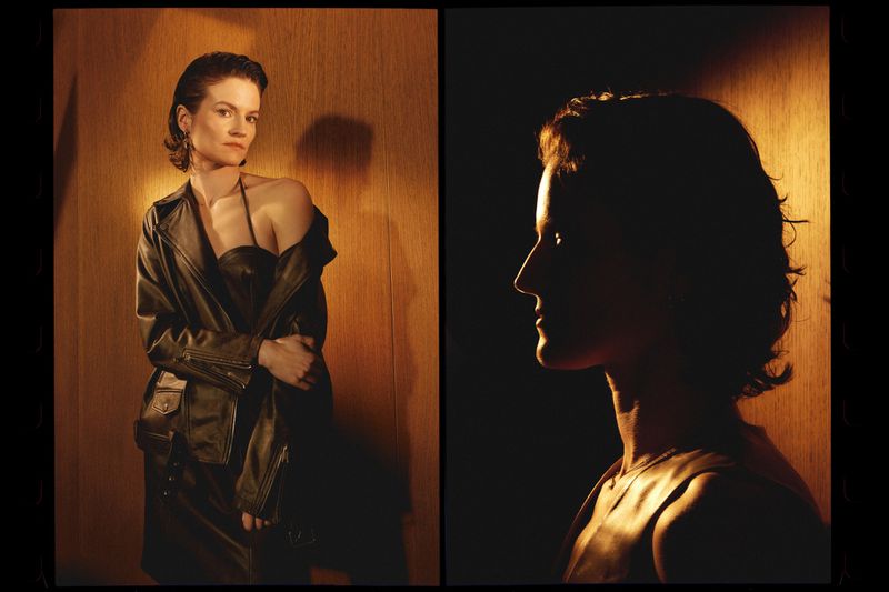 Portrait of an adult woman in a black leather jacket and dress, against a wood-paneled wall; a second profile view is silhouetted.