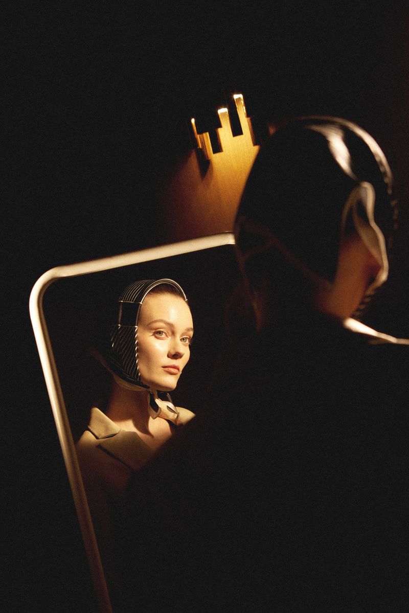 Indoor shot featuring a woman with light skin wearing a headscarf, posing with a neutral facial expression, reflected in a mirror.