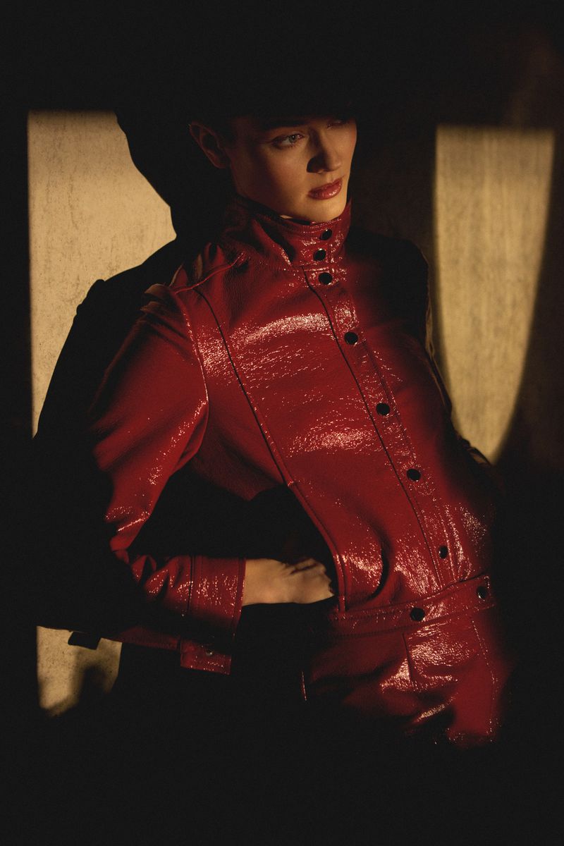 A woman in a red patent leather jacket poses in a dimly lit space with light patterns. Fashion, portrait, studio lighting.