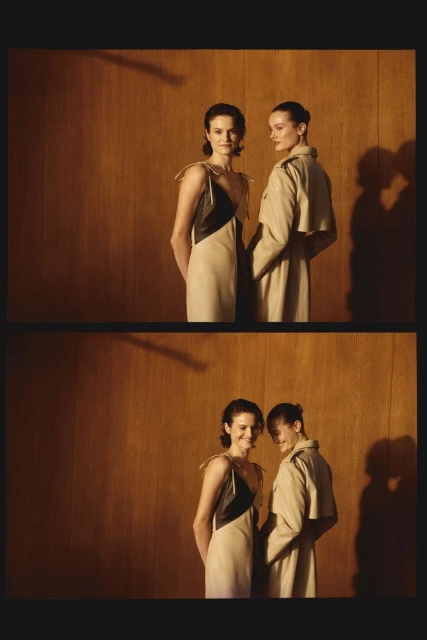 Two adult women pose in a photo studio with a wood paneled background. One wears a dress; the other wears a trench coat.